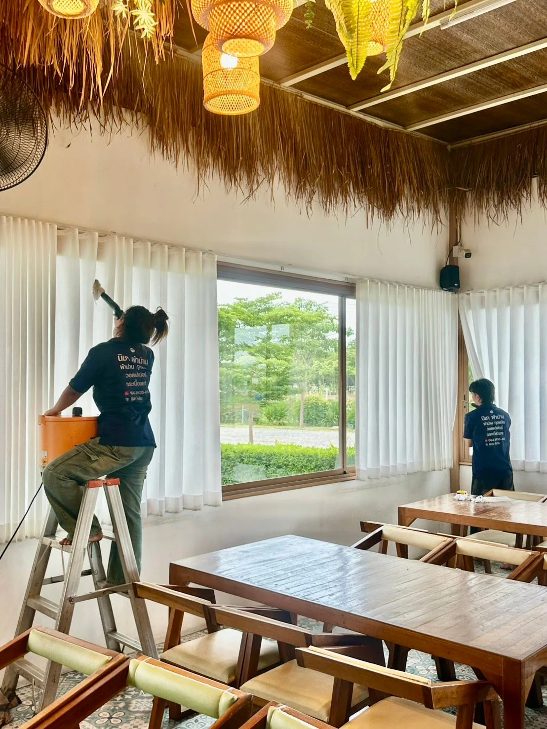 ช่างเก็บรายละเอียดม่านขาวรอบร้านอาหาร เห็นโต๊ะไม้และวิวสวนด้านนอก